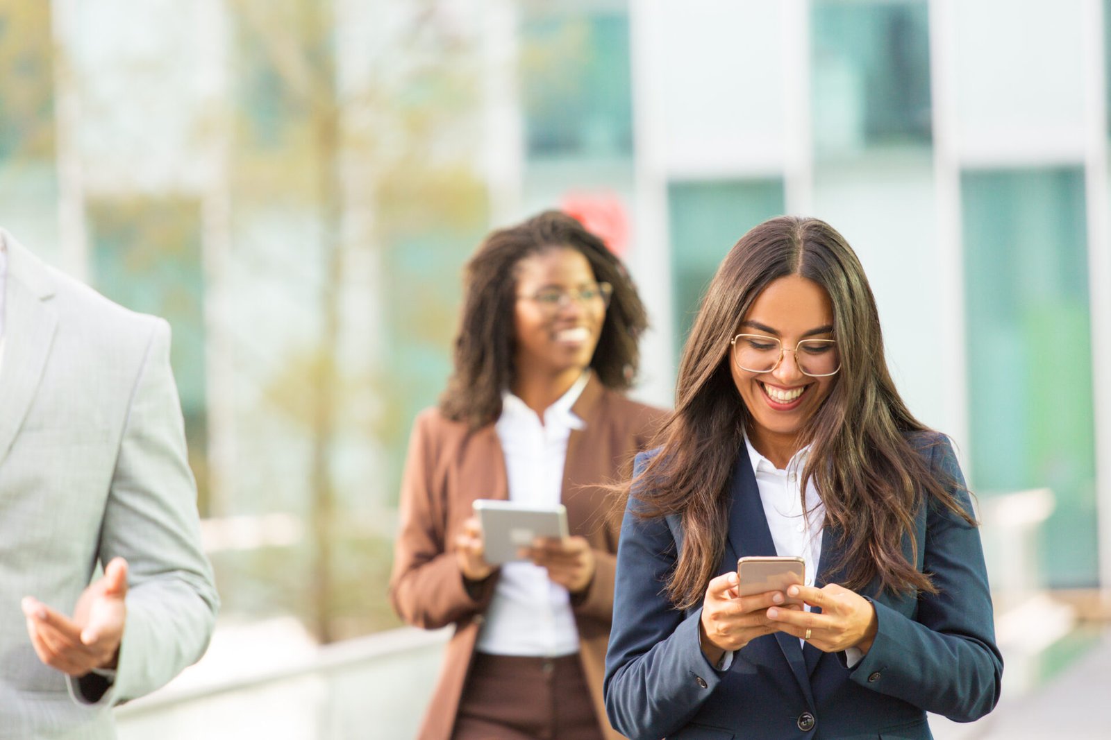 Happy Latin office employee texting message while going down city street. Young businesswoman using mobile phone, smiling at screen, woman with tablet walking behind her. Business area concept